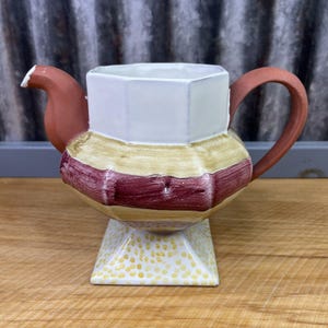 May include: A handcrafted ceramic teapot with a square white top, a bulbous body with yellow and burgundy stripes, and a square base with yellow polka dots. The spout and handle are a reddish-brown color.