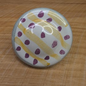 May include: A round ceramic cabinet knob with a white base featuring diagonal yellow brushstroke stripes and scattered purple polka dots. The knob has a textured, light gray border around the edge, providing a distinct contrast to the center.
