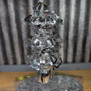 May include: A vintage silver Christmas decoration with a tiered design and intricate details. The decoration has a reflective surface and is suspended by a gold string. The base is a matching silver color. The words "Christmas Decoration" are visible on the packaging.