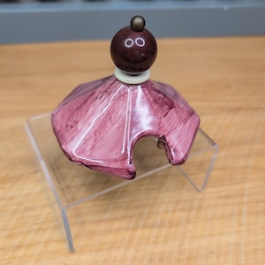 May include: A decorative bottle stopper with a maroon-colored, spherical top and a matching base. The base is a shade of pink with a textured, layered design. The stopper is displayed on a clear acrylic stand.
