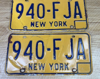 May include: Two vintage New York license plates. The plates are yellow with blue lettering and numbers. Each plate reads "940-FJA" and "NEW YORK". The plates show signs of wear and tear, with some rust and discoloration.