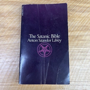 May include: A black book with the title "The Satanic Bible" by Anton Szandor LaVey. A pink pentagram with a goat's head inside is featured on the cover.