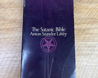 May include: A black book with the title "The Satanic Bible" by Anton Szandor LaVey. A pink pentagram with a goat's head inside is featured on the cover.
