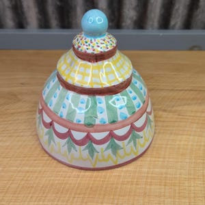 May include: A colorful ceramic jar with a lid, resembling a tiered cake. The jar features a light blue knob, yellow checkered, green striped, and floral patterns. The base has a scalloped edge with red and green accents.
