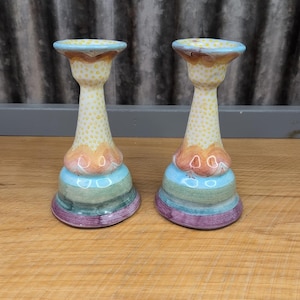 May include: Two colorful ceramic candle holders. Each has a flared base with horizontal stripes in shades of purple, green, and blue. The middle section is a light yellow with small dots. The top is a light blue with a brown rim.