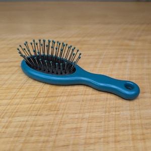 May include: A teal-colored, oval-shaped hairbrush with a handle and a hole for hanging. The brush has black bristles with teal-colored tips. The brush is made of plastic and is designed for detangling and styling hair.