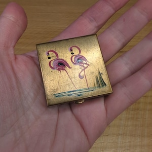 May include: A square gold-toned metal compact case featuring a painted illustration of two pink flamingos standing in blue water. The surface has a distressed, weathered texture, and the design includes small green reeds near the bottom right corner.