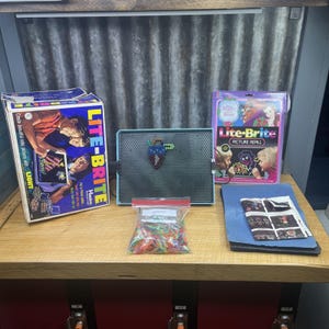 May include: A vintage Lite-Brite toy set with a light-up screen, a box, and a picture refill pack. The box features illustrations of children creating colorful pictures. A bag of colored pegs and a blue paper with designs are also present.