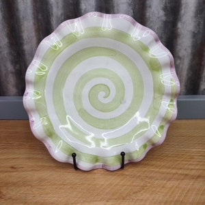 May include: A white ceramic bowl with a green and white spiral design and a pink ruffled edge. The bowl is on a black stand.