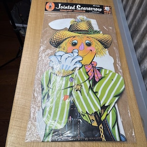 May include: A vintage, jointed cardboard cutout of a scarecrow with a green and white striped shirt, a straw hat, and a friendly face. The cutout is 32 inches tall and is completely assembled. The packaging says "Jointed Scarecrow" and "Completely Assembled".