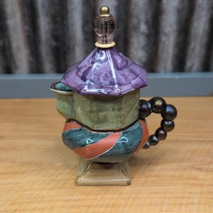 May include: A decorative ceramic teapot with a purple lid and a handle made of dark, round beads. The body of the teapot is green and blue with red stripes. The teapot sits on a clear, square base.