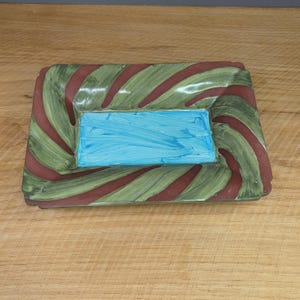 May include: Rectangular ceramic serving dish with a blue center and a green and maroon striped border. The dish has a slightly raised edge and is set on a wooden surface.