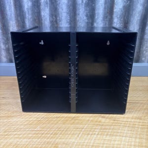 May include: A black plastic Case Logic media storage rack featuring two vertical compartments with multiple horizontal slots for organizing discs. The unit has a central divider with the brand name embossed vertically and keyhole slots on the back.