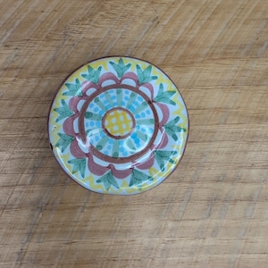 May include: A small, round, decorative plate with a colorful floral design. The plate features a yellow border, a white background, and a central pattern of green leaves, pink petals, and blue dots. The plate is approximately 4 inches in diameter.