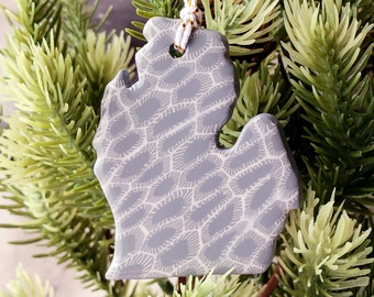 May include: A gray ceramic ornament shaped like the state of Michigan with a white patterned design. The ornament is hanging from a white and gold string.
