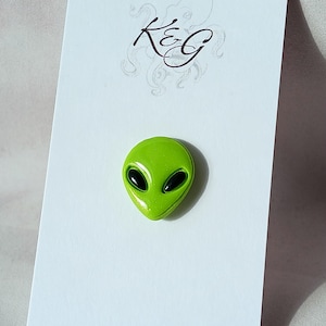 May include: A lime green alien head pin with black eyes is displayed on a white card. The card has a handwritten logo in black ink at the top. The pin is a small, novelty item.