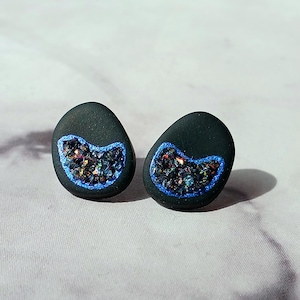 May include: A pair of black oval-shaped stud earrings with a blue outline and a crescent-shaped section filled with iridescent glitter.