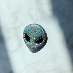 May include: Glittery alien head with large black oval eyes. The alien head is silver with a rainbow glitter effect. The alien head is a unique and eye-catching design.