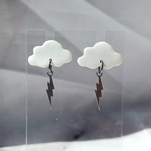 May include: A pair of white cloud earrings with silver lightning bolt charms. The earrings are displayed on a clear acrylic stand.