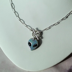 May include: A silver-toned necklace featuring a pendant shaped like an alien head. The alien pendant has a shimmering, iridescent finish with black oval eyes. The necklace has a toggle clasp and a chain link design.