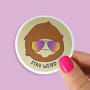 May include: A round sticker with a cartoon illustration of a brown-haired person wearing sunglasses with a gradient of purple and yellow. The text "Stay Weird" is printed below the illustration.