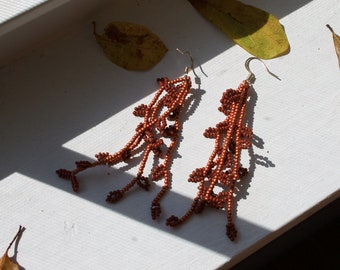 May include: A pair of copper-coloured beaded earrings. The earrings feature a cascading design with small, round beads and silver-coloured ear wires. The earrings are displayed on a white surface with dried leaves.
