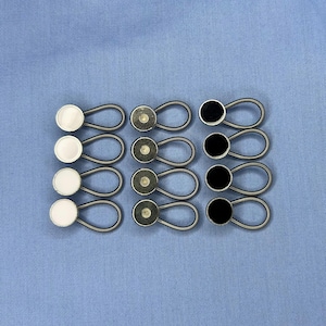 May include: A set of twelve elastic button extenders arranged in three columns. The left column features four white circular buttons, the middle column has four metallic silver-toned buttons, and the right column contains four black circular buttons.
