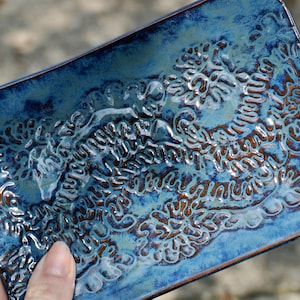 May include: A rectangular ceramic tray featuring a glossy blue glaze with dark brown, textured, raised patterns across the surface. The tray has rounded corners and a deep, reflective finish that highlights the intricate, swirling, organic designs.