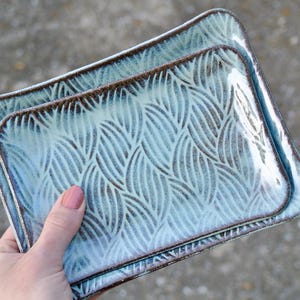 May include: Two rectangular ceramic trays with a light blue and white glaze featuring a dark brown leaf-like pattern. The trays have slightly raised edges and a glossy finish, with the smaller tray nested inside the larger one.