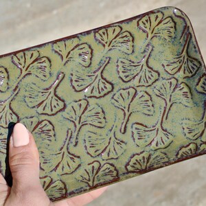 May include: A rectangular ceramic tray featuring a glossy olive green glaze with a repeating embossed pattern of dark brown ginkgo leaves. The edges of the tray are finished with a dark brown rim, highlighting the textured leaf impressions.