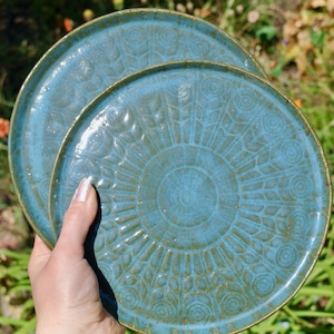 May include: Two round ceramic plates with a glossy blue glaze and a textured, radial pattern featuring concentric circles and leaf-like motifs. The plates have a slightly raised rim and a dark, earthy brown edge that contrasts with the blue surface.