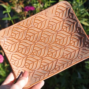 May include: A rectangular ceramic tray with a terracotta-colored glaze featuring a repeating embossed leaf pattern. The surface has a smooth finish with darker brown accents highlighting the recessed lines of the leaf design.