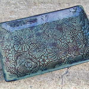 May include: A rectangular ceramic tray featuring a glossy finish with a deep teal and green glaze. The surface is decorated with a repeating dark brown line-art pattern of various flowers and petals covering the entire interior area.