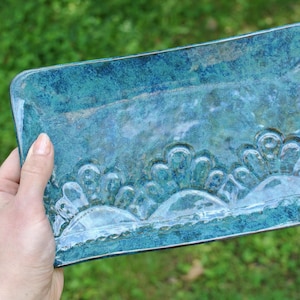 May include: A rectangular ceramic tray with a mottled teal and blue glaze. The surface features a raised, embossed pattern of three repeating semi-circular fan shapes along the bottom edge, creating a textured, glossy finish on the pottery.