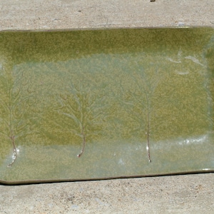 May include: A rectangular ceramic tray with a glossy olive green glaze and a smooth finish. The surface features four embossed tree silhouettes arranged in a row, with subtle variations in the green color tones across the flat, shallow base.