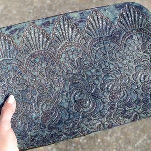 May include: A rectangular ceramic tray with rounded corners, featuring a glossy teal and dark blue glaze. The surface is decorated with a raised, intricate lace-like pattern that covers the entire top, creating a textured and detailed appearance.