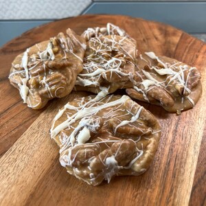 Puede incluir: Una tabla de madera presenta varias pralin&eacute;s de nuez pecana. Las pralin&eacute;s son de color marr&oacute;n claro, con trozos de nuez pecana y un glaseado blanco. El fondo muestra una pared de azulejos azules y blancos.