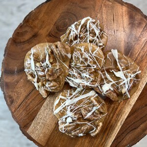 Puede incluir: Vista superior de cinco pralin&eacute;s de nuez pecana sobre una tabla de madera. Los pralin&eacute;s son de color marr&oacute;n claro con trozos de nuez pecana visibles y est&aacute;n rociados con glaseado blanco. La tabla de madera tiene vetas naturales y es circular.