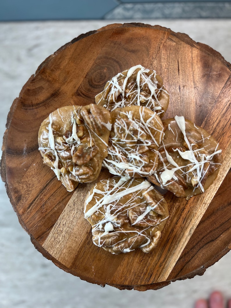 Puede incluir: Vista superior de cinco pralin&eacute;s de nuez pecana sobre una tabla de madera. Los pralin&eacute;s son de color marr&oacute;n claro con trozos de nuez pecana visibles y est&aacute;n rociados con glaseado blanco. La tabla de madera tiene vetas naturales y es circular.