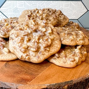 Puede incluir: Una pila de pralin&eacute;s de nuez pecana sobre una tabla de madera. Los pralin&eacute;s tienen una capa brillante de color marr&oacute;n claro y est&aacute;n cubiertos de trozos de nuez pecana. La tabla de madera tiene un borde natural y r&uacute;stico.