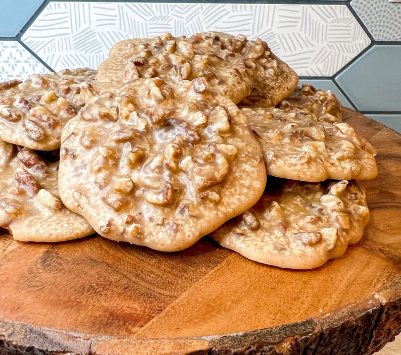 Puede incluir: Una pila de pralin&eacute;s de nuez pecana sobre una tabla de madera. Los pralin&eacute;s tienen una capa brillante de color marr&oacute;n claro y est&aacute;n cubiertos de trozos de nuez pecana. La tabla de madera tiene un borde natural y r&uacute;stico.