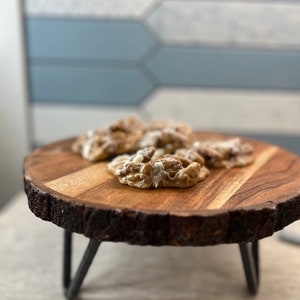 Puede incluir: Una bandeja de madera con borde natural y patas de metal, que muestra varios productos horneados con glaseado blanco. La bandeja es redonda y est&aacute; hecha de madera de color marr&oacute;n claro. El fondo presenta una pared de azulejos azules y blancos.