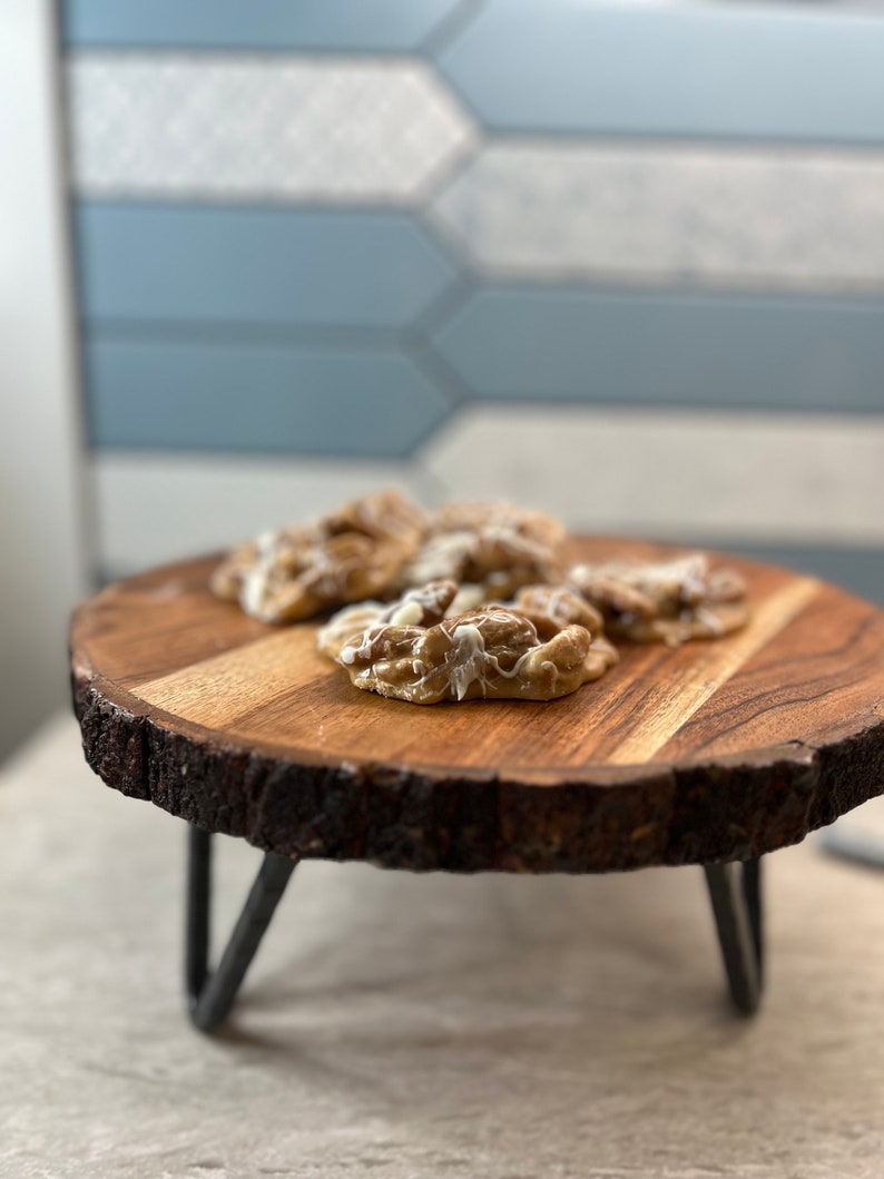 Puede incluir: Una bandeja de madera con borde natural y patas de metal, que muestra varios productos horneados con glaseado blanco. La bandeja es redonda y est&aacute; hecha de madera de color marr&oacute;n claro. El fondo presenta una pared de azulejos azules y blancos.