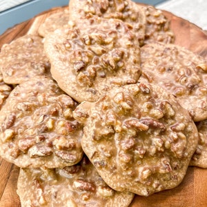 Puede incluir: Una fuente de madera contiene una pila de pralin&eacute;s de nuez. Los caramelos tienen una capa brillante de color marr&oacute;n claro y est&aacute;n salpicados de trozos de nuez. Los pralin&eacute;s est&aacute;n dispuestos de forma ligeramente superpuesta, mostrando su forma redonda y superficie texturizada.