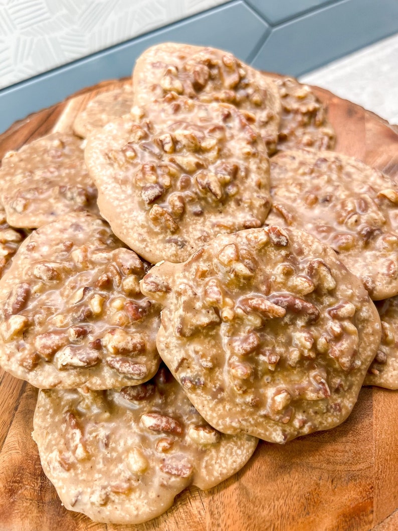 Puede incluir: Una fuente de madera contiene una pila de pralin&eacute;s de nuez. Los caramelos tienen una capa brillante de color marr&oacute;n claro y est&aacute;n salpicados de trozos de nuez. Los pralin&eacute;s est&aacute;n dispuestos de forma ligeramente superpuesta, mostrando su forma redonda y superficie texturizada.