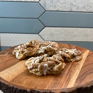 Puede incluir: Varias pralin&eacute;s de nueces pecanas, rociadas con glaseado blanco, se encuentran sobre una tabla redonda de madera. Las pralin&eacute;s son de color marr&oacute;n claro con nueces pecanas visibles. La tabla tiene un borde natural, sobre un fondo de azulejos azules y blancos.