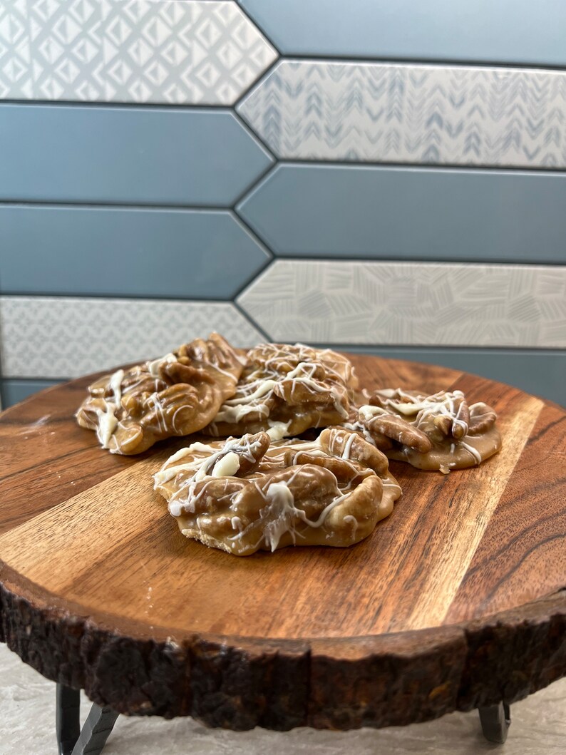 Puede incluir: Varias pralin&eacute;s de nueces pecanas, rociadas con glaseado blanco, se encuentran sobre una tabla redonda de madera. Las pralin&eacute;s son de color marr&oacute;n claro con nueces pecanas visibles. La tabla tiene un borde natural, sobre un fondo de azulejos azules y blancos.