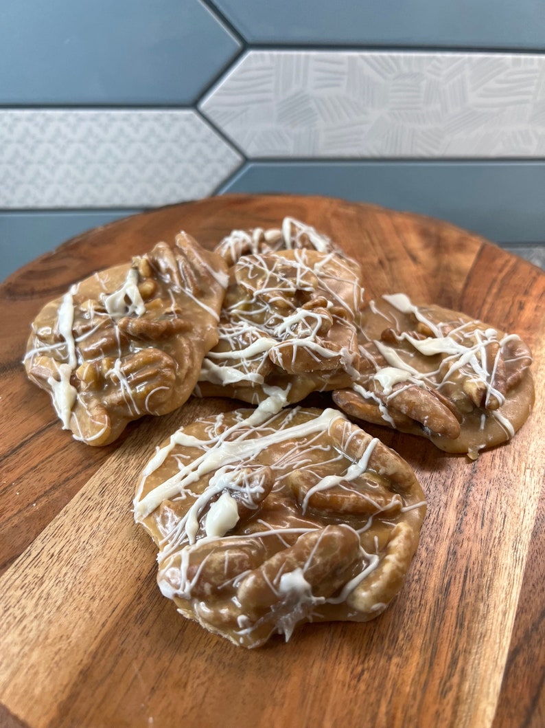 Puede incluir: Una tabla de madera presenta varias pralin&eacute;s de nuez pecana. Las pralin&eacute;s son de color marr&oacute;n claro, con trozos de nuez pecana y un glaseado blanco. El fondo muestra una pared de azulejos azules y blancos.