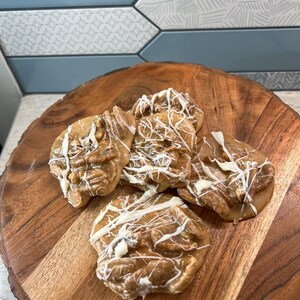 Puede incluir: Varias pralin&eacute;s de nueces pecanas con un glaseado de chocolate blanco sobre una tabla redonda de madera. Las pralin&eacute;s son de color marr&oacute;n claro con nueces pecanas visibles y un acabado brillante. El fondo presenta un patr&oacute;n de baldosas decorativas.