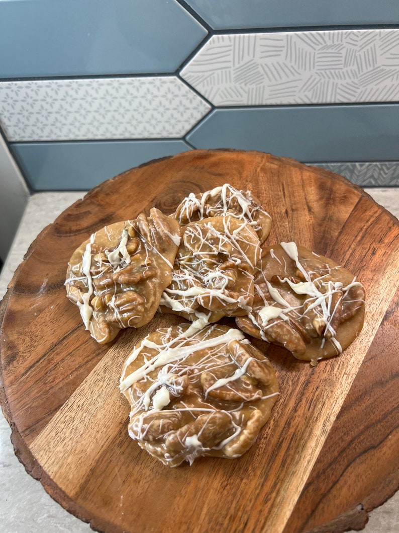 Puede incluir: Varias pralin&eacute;s de nueces pecanas con un glaseado de chocolate blanco sobre una tabla redonda de madera. Las pralin&eacute;s son de color marr&oacute;n claro con nueces pecanas visibles y un acabado brillante. El fondo presenta un patr&oacute;n de baldosas decorativas.
